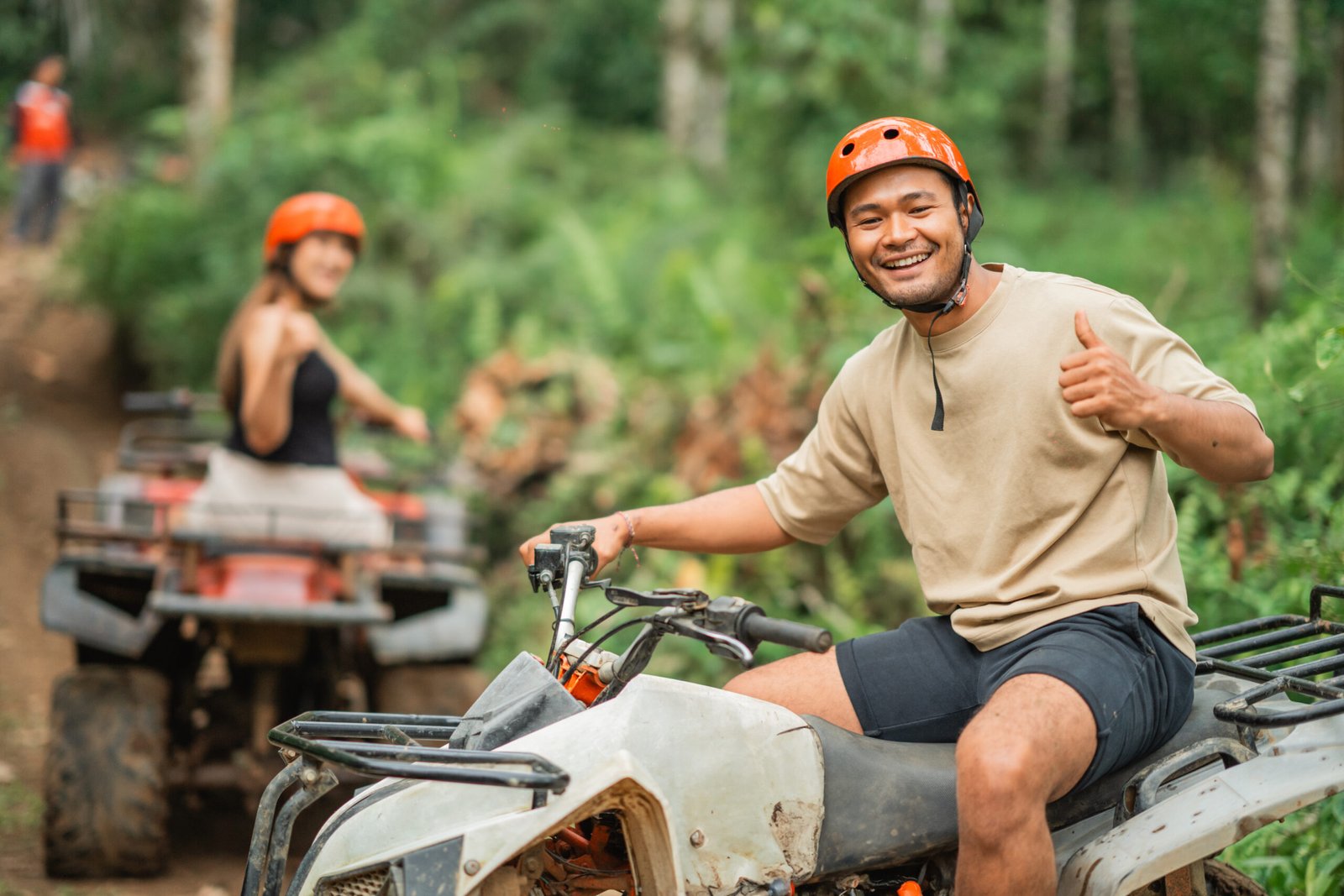 handsome asian man smiling to the camera with thumb up while riding the atv with his beautiful partner at the background